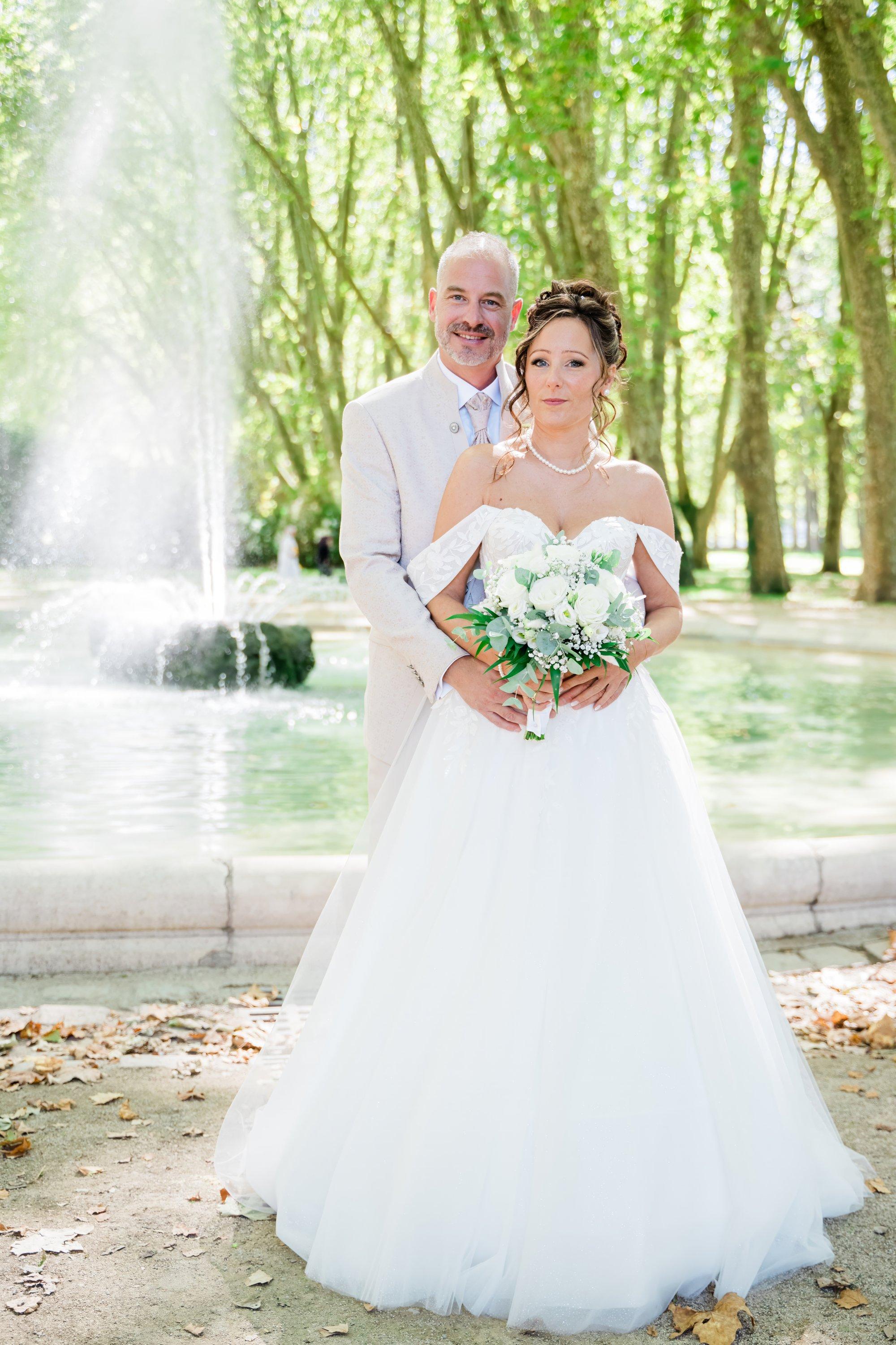 Mariés posant devant la fontaine du parc avec un bouquet blanc