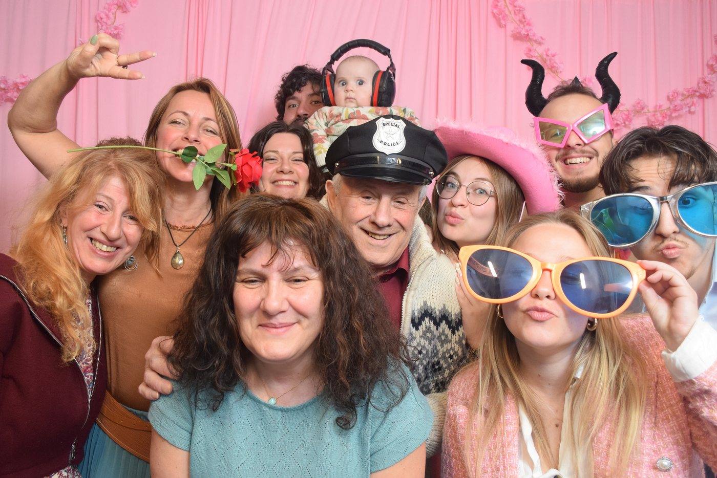 Famille posant avec des accessoires devant le fond rose fleuri du photobooth lors d'un baptême