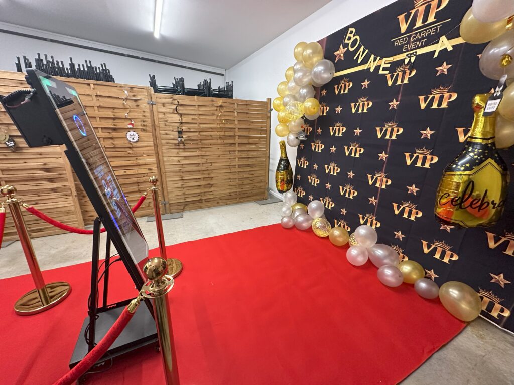 Soirée Photobooth, Tapis rouge - potelets dorés Espace Photobooth avec tapis rouge, potelets dorés et borne photo interactive. Décor VIP avec ballons dorés et blancs, fond noir et or, et table d’accessoires pour une ambiance élégante et festive.