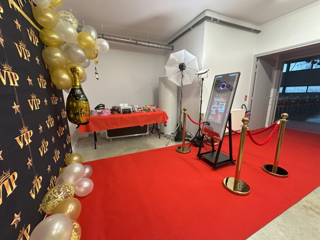 Soirée Photobooth, Tapis roupe et potelets dorés Espace Photobooth VIP avec tapis rouge, potelets dorés et borne photo interactive. Décoration festive avec ballons dorés et blancs, arrière-plan VIP noir et or, accessoires et table de friandises en arrière-plan.